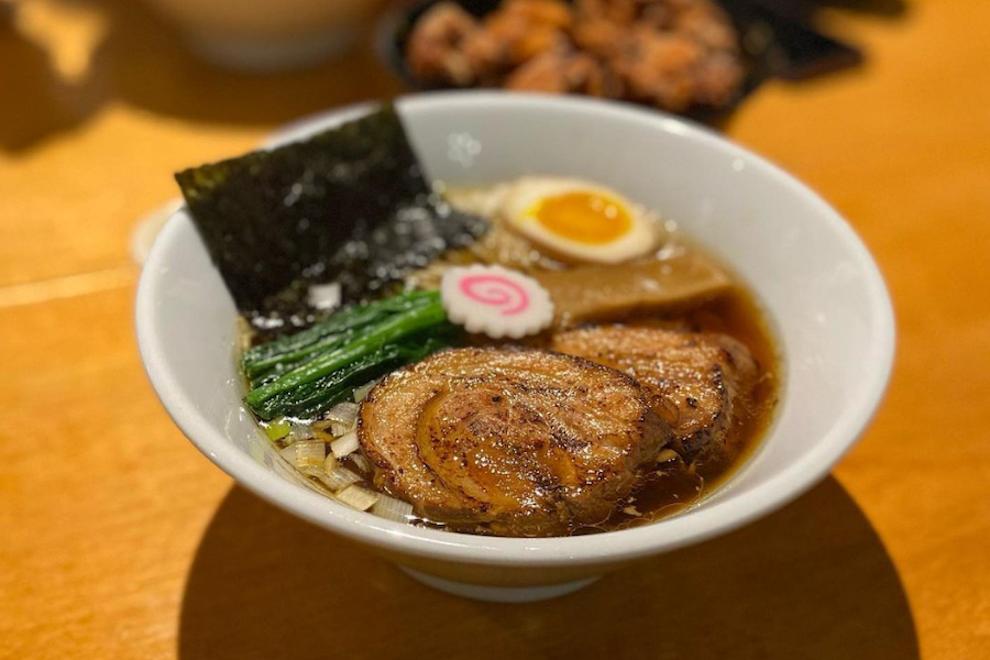 A bowl of ramen with slices of roasted pork, seaweed, boiled egg, narutomaki, and green vegetables on a wooden table.