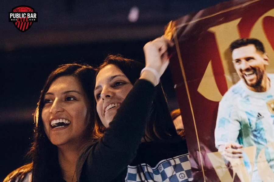 Two women smiling and holding a poster featuring Lionel Messi at Public Bar Live.