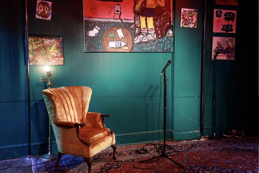 A cozy stage setup at a venue featuring a vintage armchair, a microphone on a stand, and artwork displayed on the green-painted walls.