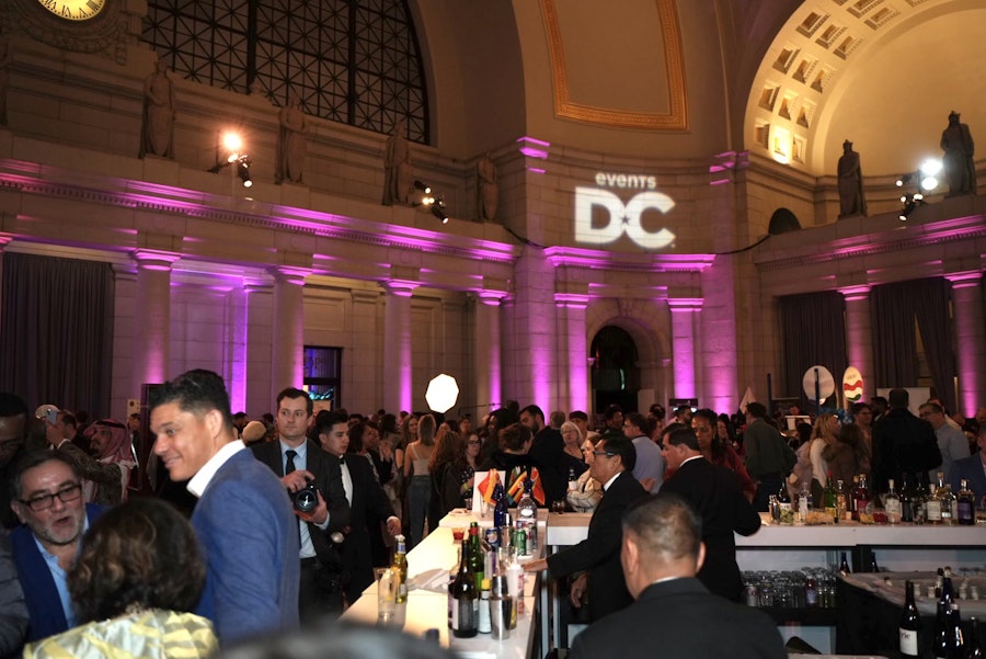 A lively event taking place in a grand hall with high ceilings and arched windows, illuminated with purple lighting. The "Events DC" logo is projected onto the wall above the crowd. People are gathered around a bar area, engaging in conversation, with some dressed in formal attire. The atmosphere appears festive and vibrant.