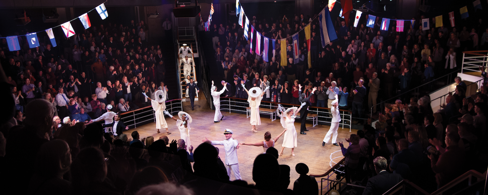 A lively scene from "Anything Goes" with actors in 1930s-style costumes on a ship deck set. The audience claps enthusiastically, surrounded by colorful flags hanging overhead.