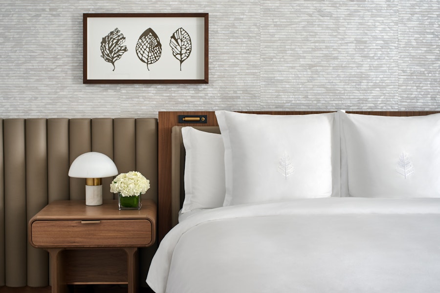 A minimalist bedroom with crisp white linens, a wooden headboard, and a bedside table with a small lamp and vase. The wall above the bed features a simple framed artwork.