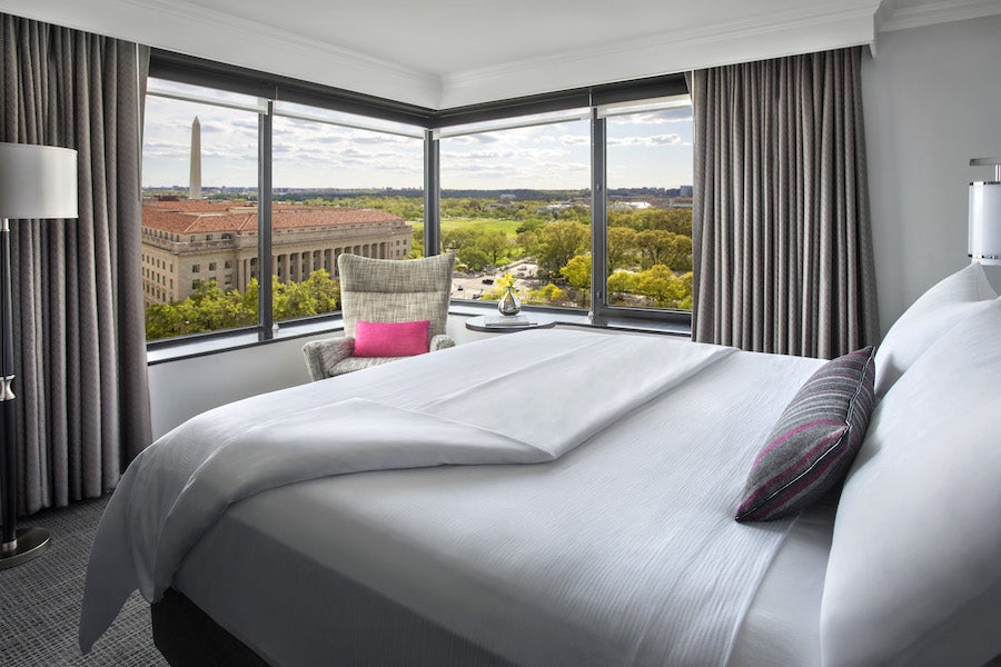A luxurious hotel room with a cozy bed and a corner view of Washington DC, featuring the Washington Monument and lush greenery outside the large windows.