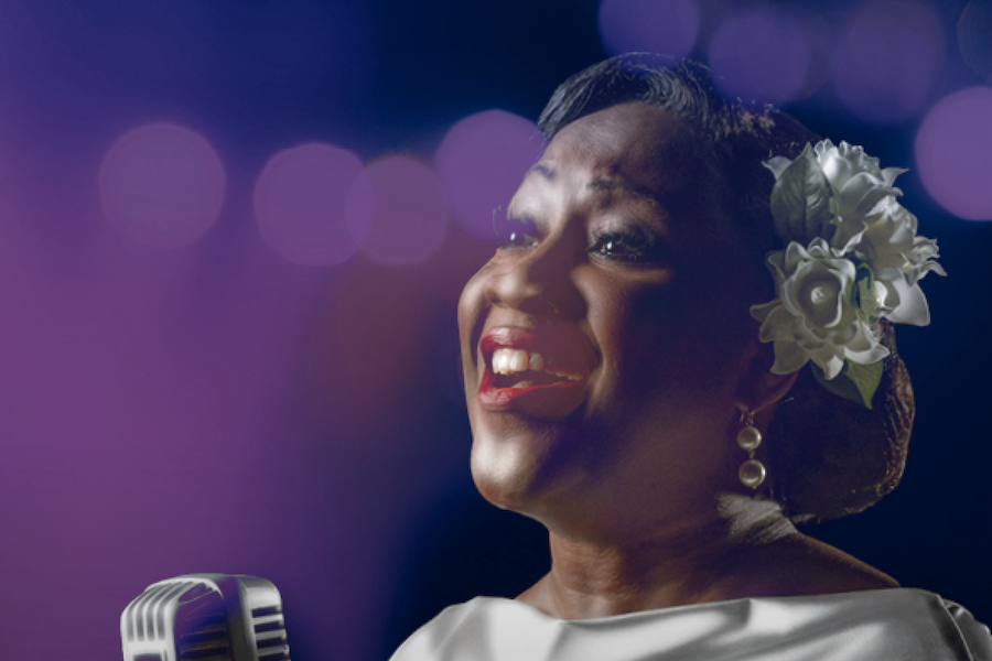 A woman sings passionately into a vintage microphone, wearing a white flower in her hair and earrings, with a backdrop of blurred purple lights.