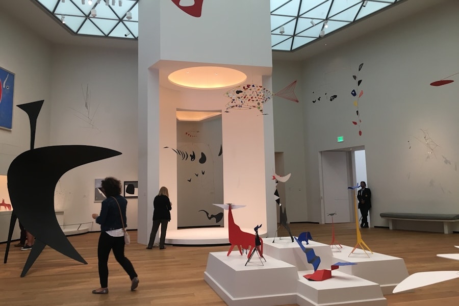 Visitors explore the Calder Room at the National Gallery of Art, showcasing colorful and abstract mobile sculptures by Alexander Calder, suspended from the ceiling and displayed on pedestals.