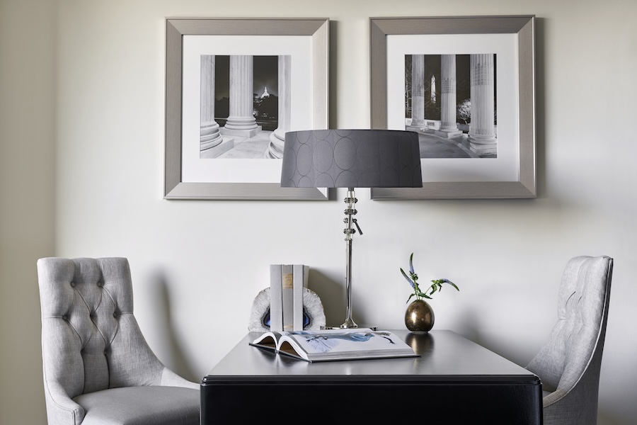 Small desk area with two gray upholstered chairs, a modern lamp, and two black and white framed photos of architectural columns on the wall.