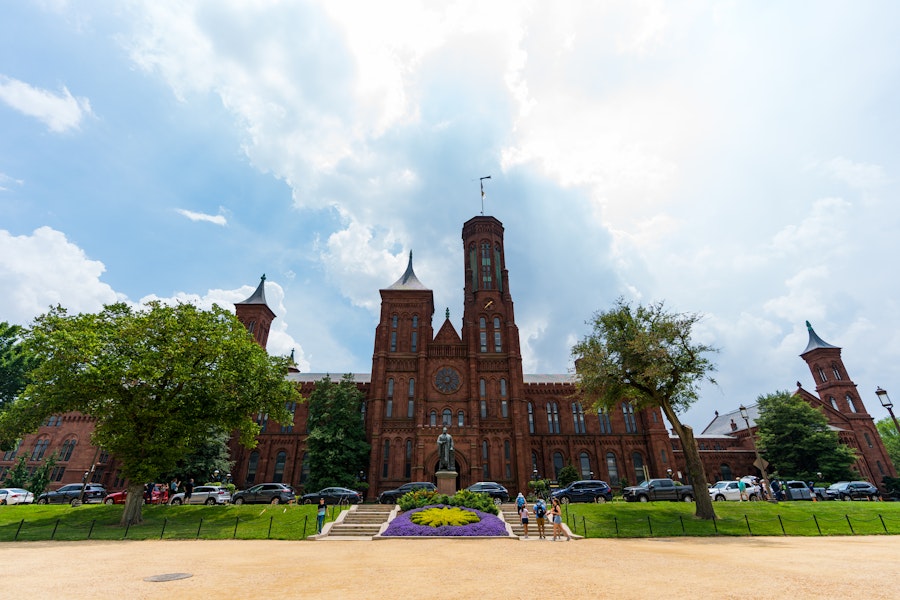 Exterior view of the Smithsonian Castle on the National Mall, with a statue of Joseph Henry in front and people walking nearby under a partly cloudy sky.