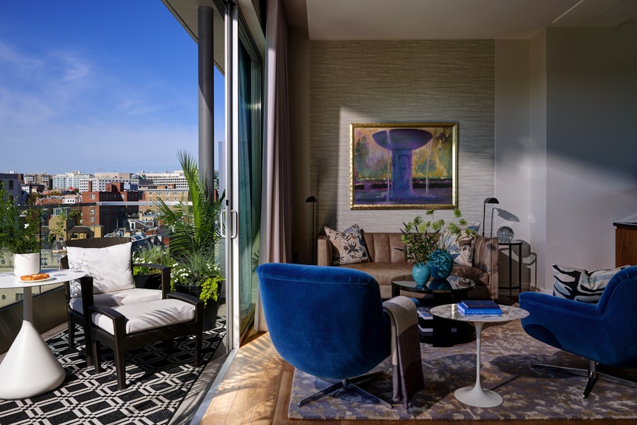 A bright living room with blue velvet chairs, a comfortable couch, and a sliding glass door that opens to a balcony with a view of the city. A large, colorful painting hangs on the wall.