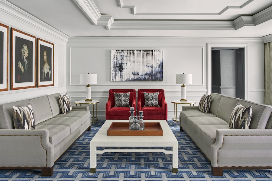 An elegant living room with two gray sofas and two red armchairs arranged around a coffee table. The walls are adorned with framed portraits, and a large abstract painting hangs above the armchairs.