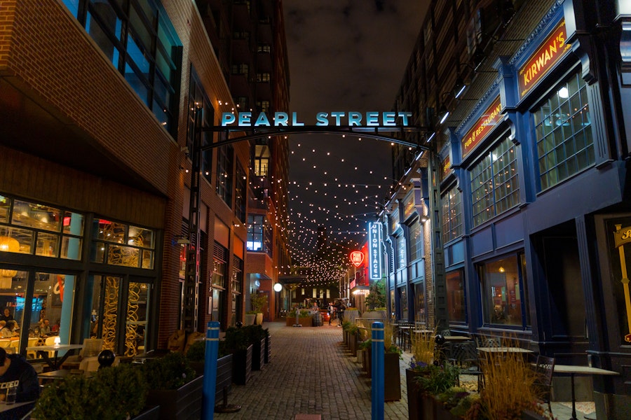 A view of Pearl Street at The Wharf in Washington, D.C., at night, with string lights hanging overhead. The street is lined with restaurants and pubs, including Kirwan's, creating a lively and inviting atmosphere. The "PEARL STREET" sign arches over the entrance to the narrow, cobblestone path.