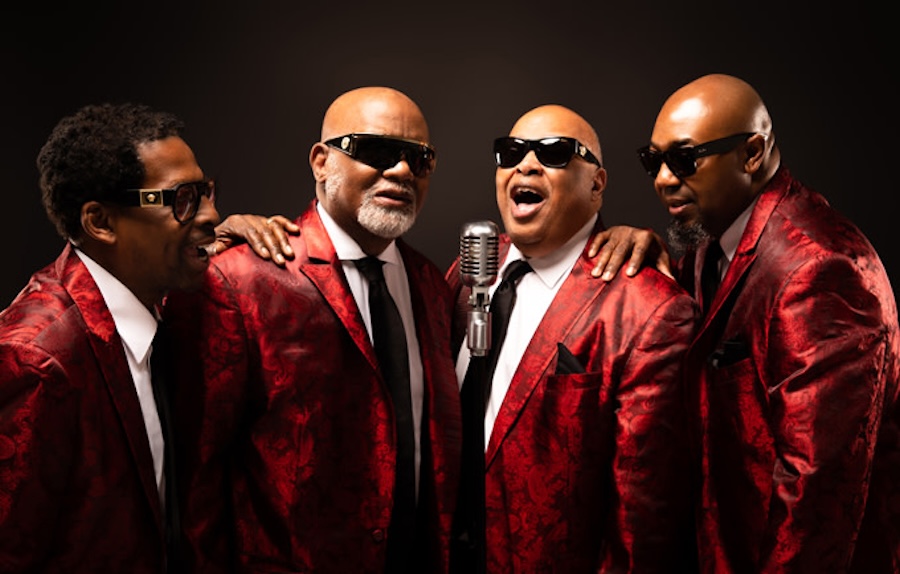 Four men, members of the Blind Boys of Alabama, are wearing matching red patterned suits and sunglasses. They stand close together around a vintage microphone, with one man singing while the others smile.