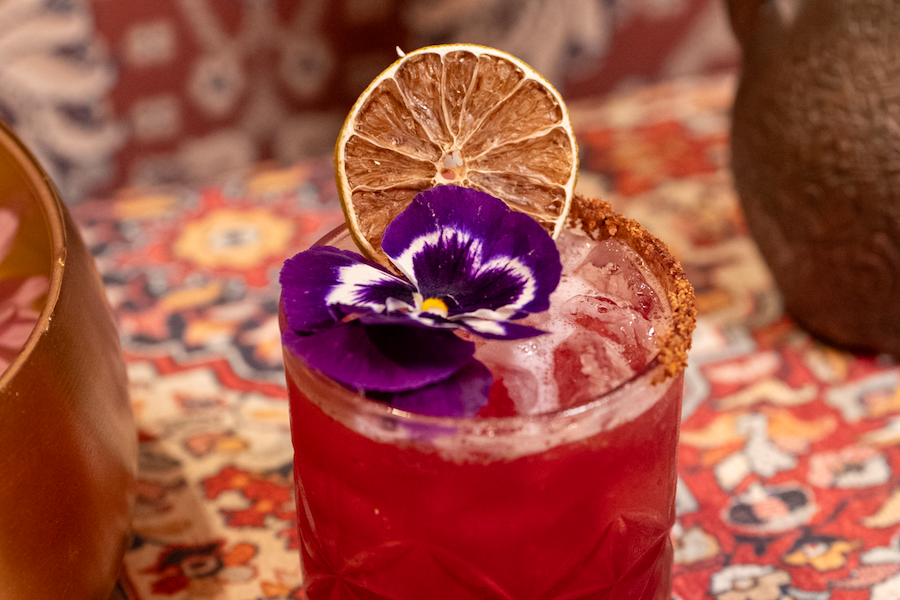 A vibrant red cocktail served in a glass with a salt rim, garnished with a dried lime slice and a purple flower. The drink is placed on a colorful patterned table.