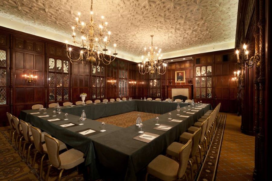 An elegant, traditional conference room at the Hay-Adams, featuring dark wood paneling, ornate chandeliers, and a U-shaped table setup under a decorative, intricate ceiling.