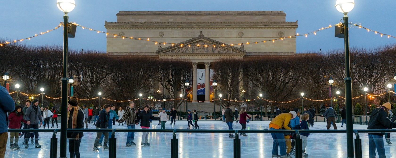 People ice skating at the National Gallery of Art Sculpture Garden rink during the evening, with string lights overhead and the museum building in the background.