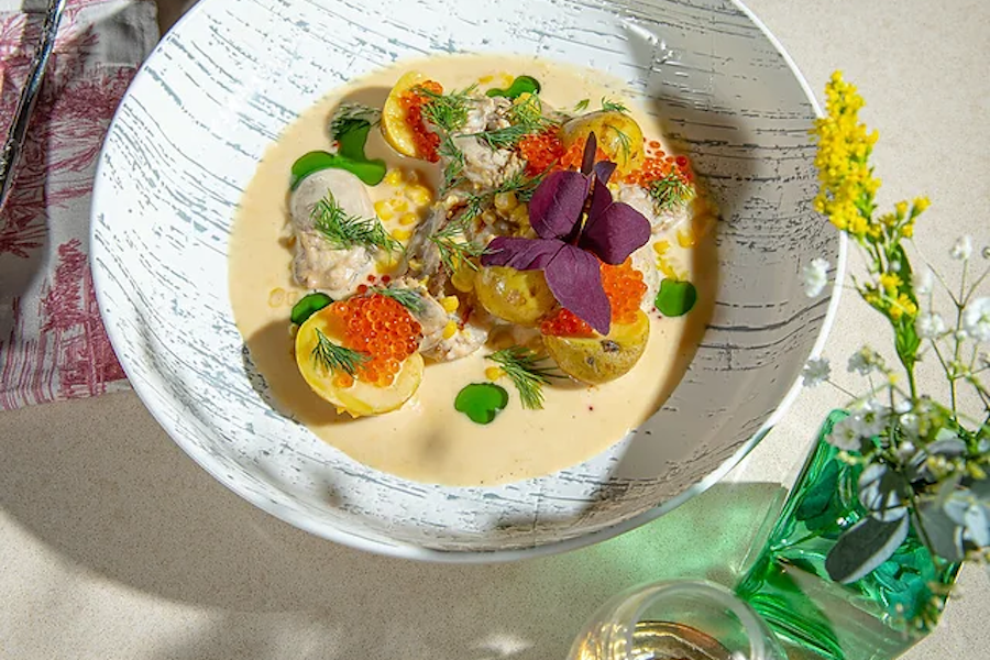 A seafood dish with potatoes, orange roe, and dill in a creamy sauce, garnished with purple microgreens. A small vase with yellow flowers sits nearby.