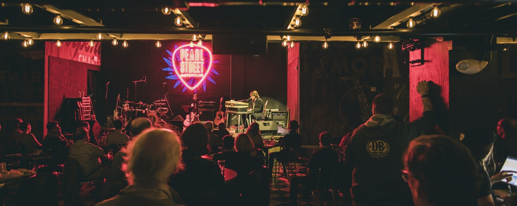 An intimate live music performance at Pearl Street Warehouse, with a neon sign in the shape of a guitar pick glowing on the back wall. The audience is seated, watching the performer on stage, with warm lighting overhead.