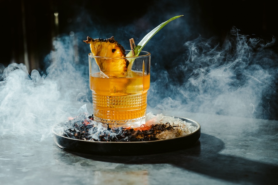 A vibrant cocktail served in a short glass, garnished with grilled pineapple and a green leaf, resting on a bed of smoking ingredients on a black plate, creating a dramatic visual effect.