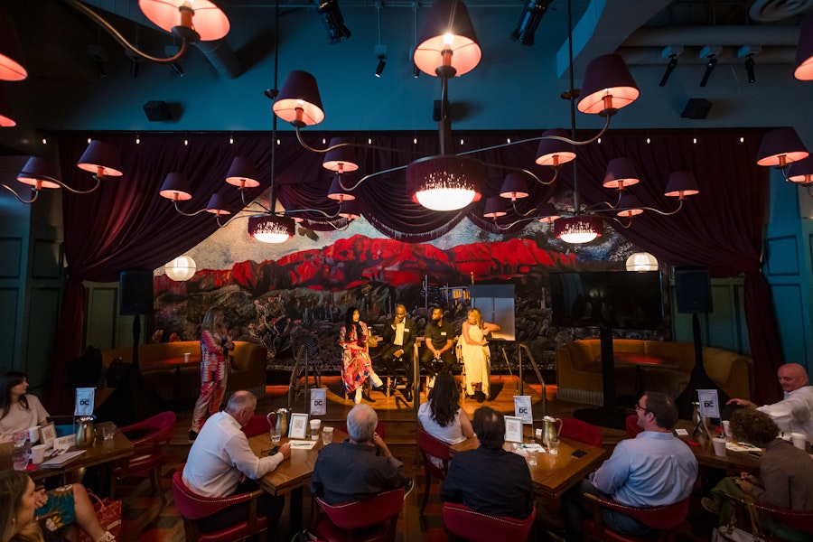 A vibrant indoor setting with a lively panel discussion taking place on a small stage. The room is adorned with dramatic red curtains, unique ceiling lights, and a colorful mural in the background, while guests sit at tables, engaged in the conversation.