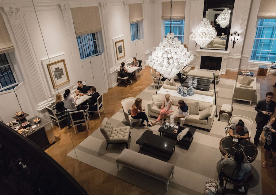 An elegant lounge area inside a historic mansion, featuring modern furnishings and a grand chandelier. People are seated in small groups, engaging in conversations or working, surrounded by comfortable chairs, sofas, and warm lighting, creating a cozy yet sophisticated atmosphere.