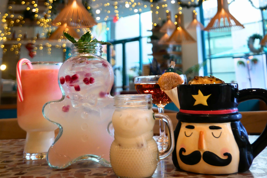 A collection of festive holiday-themed drinks in unique mugs and glasses, set against a backdrop of twinkling lights.