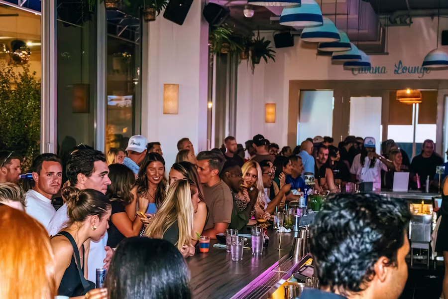 A vibrant and crowded bar scene with patrons socializing and enjoying drinks at a long counter under colorful hanging lights. The atmosphere is lively, with a mix of conversations and laughter filling the space.