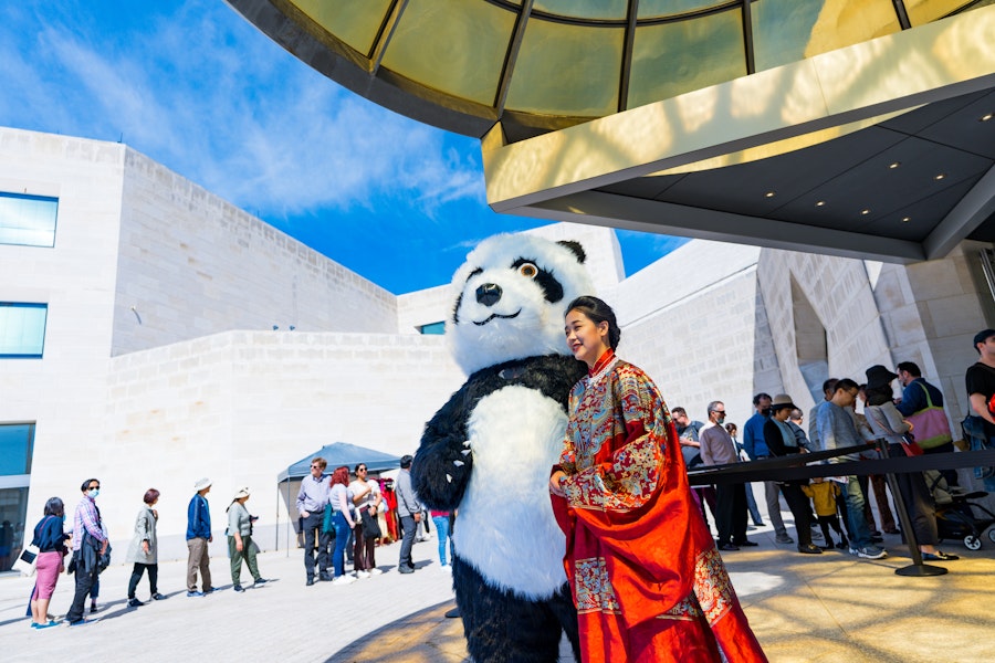 A person in a panda costume poses with a woman wearing traditional attire outside a modern building at PassportDC in Washington, DC.