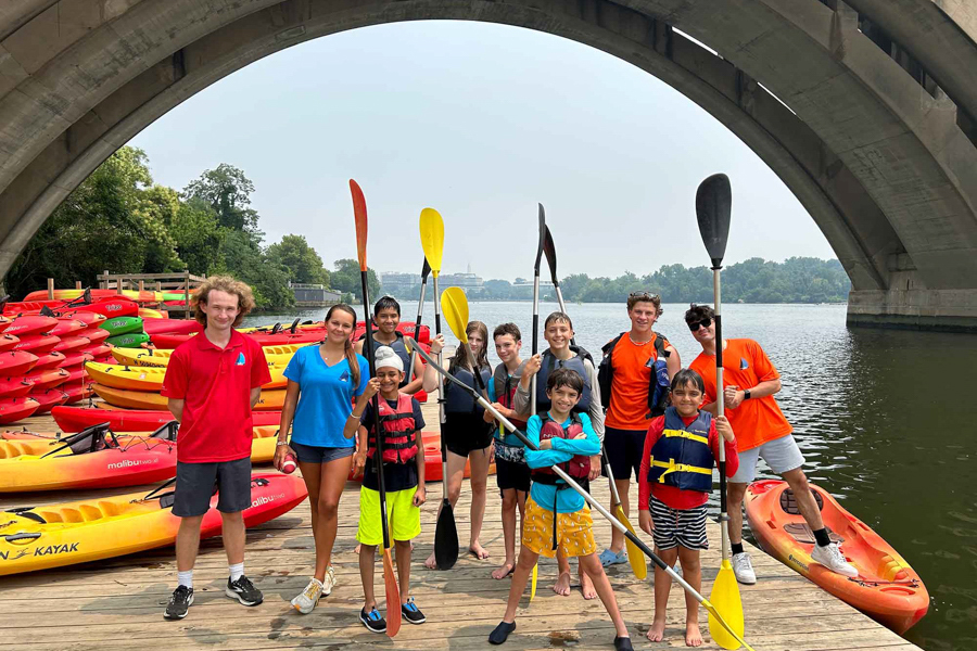 Group of kids and instructors with paddles posing on a dock surrounded by colorful kayaks under a bridge.