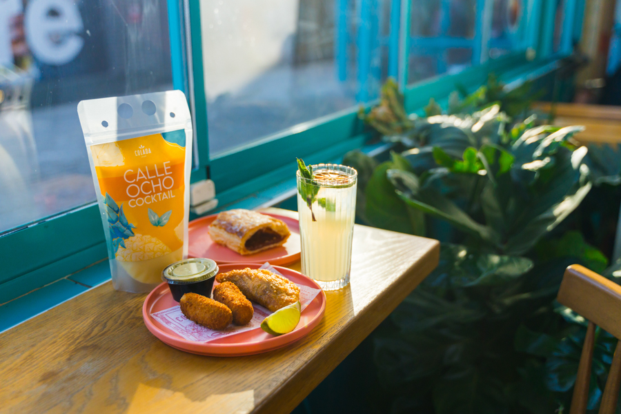 A sunlit café table displays croquetas, empanadas, a mojito, and a Calle Ocho cocktail pouch beside a teal window.