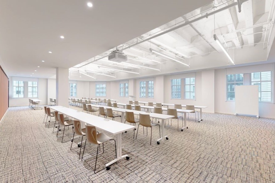 A modern, well-lit conference room with white walls, long white tables, wooden chairs, and large windows, designed for flexible and collaborative workspace needs.