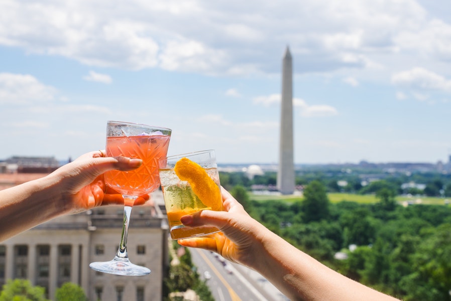 Two hands raise colorful cocktails against the backdrop of the Washington Monument from a scenic rooftop bar.