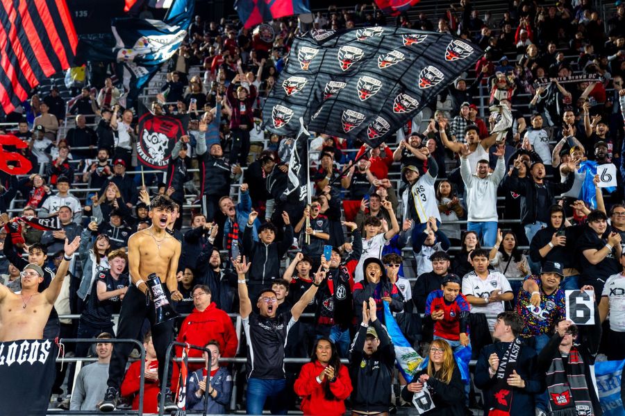 A passionate crowd of D.C. United fans cheering in the stands, waving flags and banners in black and red.