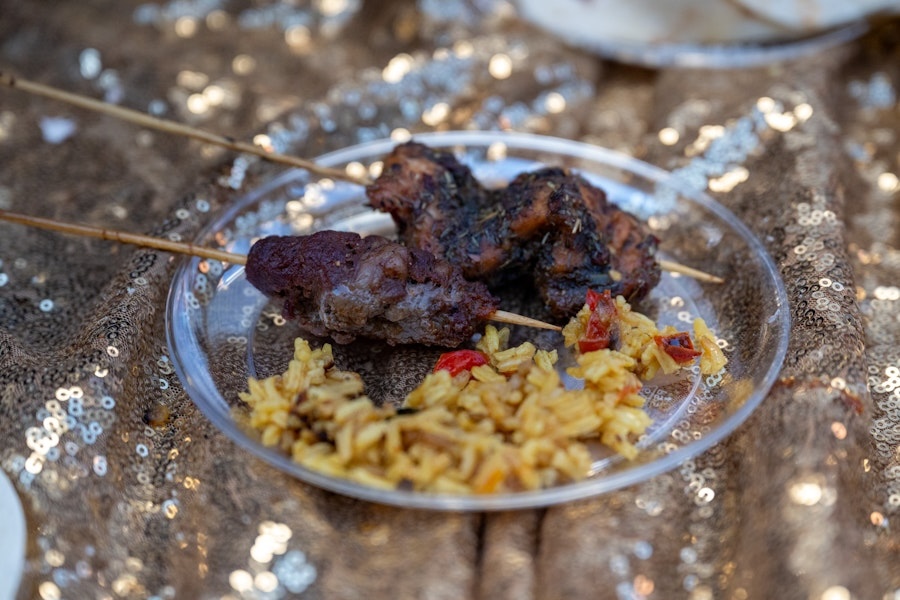 A clear plastic plate holds skewers of grilled meat alongside seasoned yellow rice with red peppers, set on a shimmering gold tablecloth.