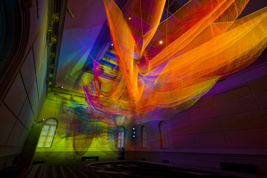 A colorful, illuminated net sculpture hangs from the ceiling, casting vibrant hues across the historic gallery walls.