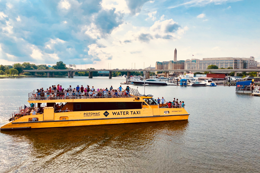 A bright yellow water taxi cruises along the Potomac River near The Wharf, with the Washington Monument visible in the background.