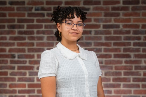 The professional headshot of Yovanna, a young Black Latina. She is wearing a white shirt and stands in front of a brick wall. 