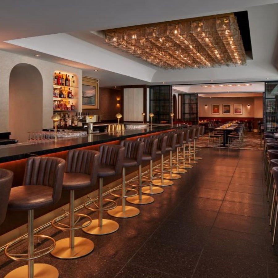 A stylish restaurant and bar with a long, sleek counter lined with leather barstools, illuminated liquor shelves, and a warmly lit dining area with modern decor in the background.