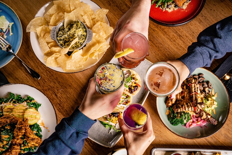 Friends toast with colorful cocktails over a table filled with tacos, spinach dip and hearty entrées at a lively restaurant.