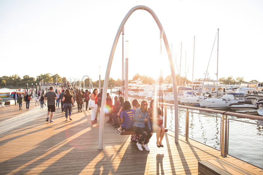 People relax on swings and stroll along the waterfront pier at The Wharf, with boats docked nearby in the golden evening light.