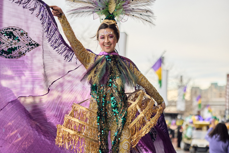 Person in a vibrant Mardi Gras costume with sequins, feathers, and a purple cape dances at an outdoor parade.