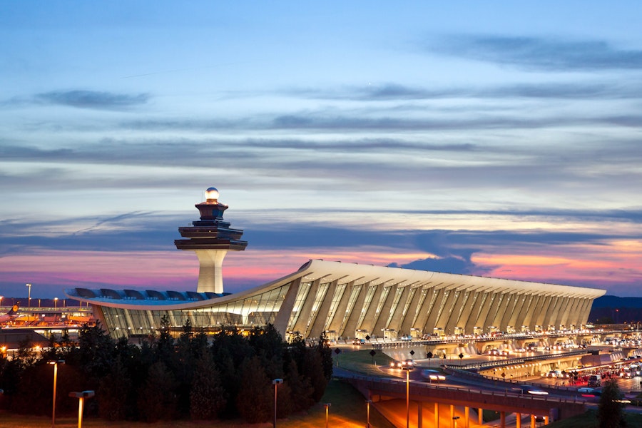 A modern airport terminal with a distinctive curved roof and control tower is illuminated at sunset, with busy traffic below.