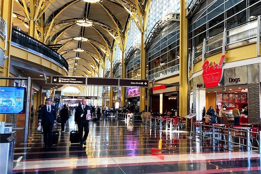 A bustling airport terminal features arched ceilings, travelers walking with luggage, and a food court including Ben's Chili Bowl.