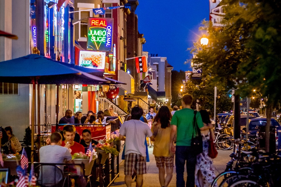 People walk down a street lit up with neon signs and busy restaurants and bars.