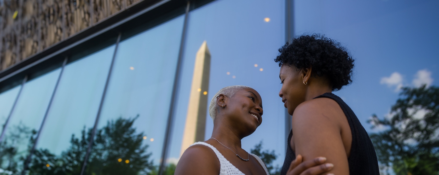 A couple shares an intimate moment in front of the Smithsonian National Museum of African American History and Culture, with the Washington Monument reflected in the glass.