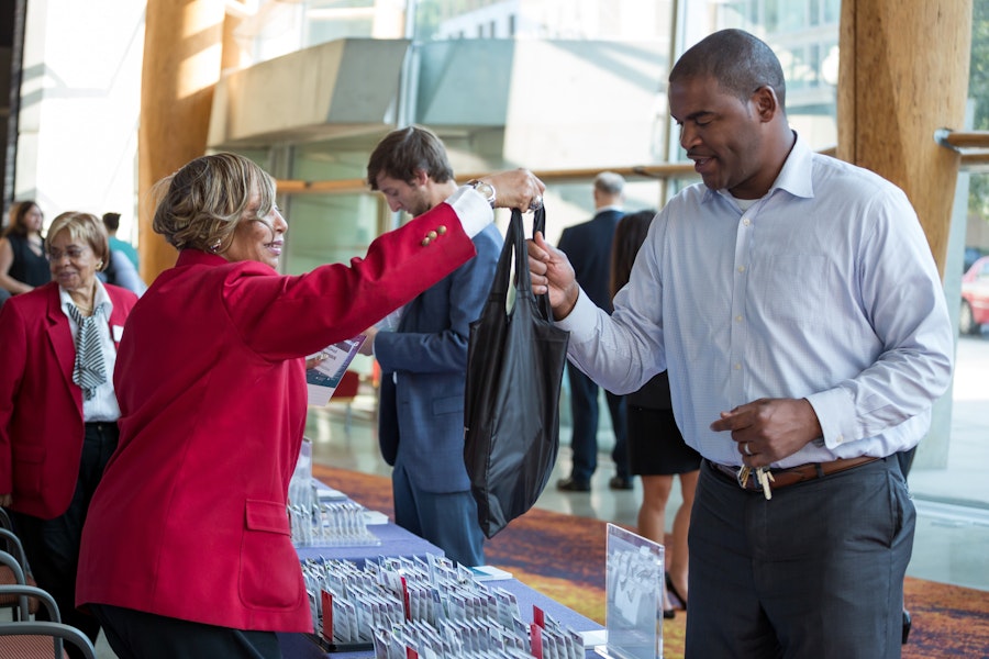 A woman in a red jacket hands a bag to a man in a white shirt at an event check-in or registration table.