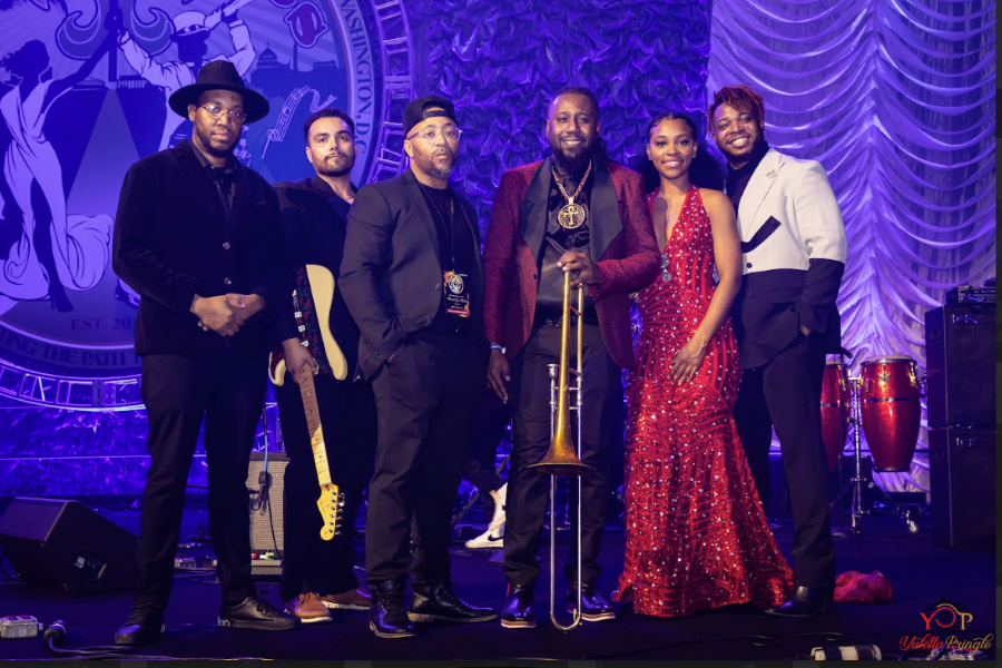 A group of musicians dressed in elegant attire pose together on stage in front of a purple backdrop.