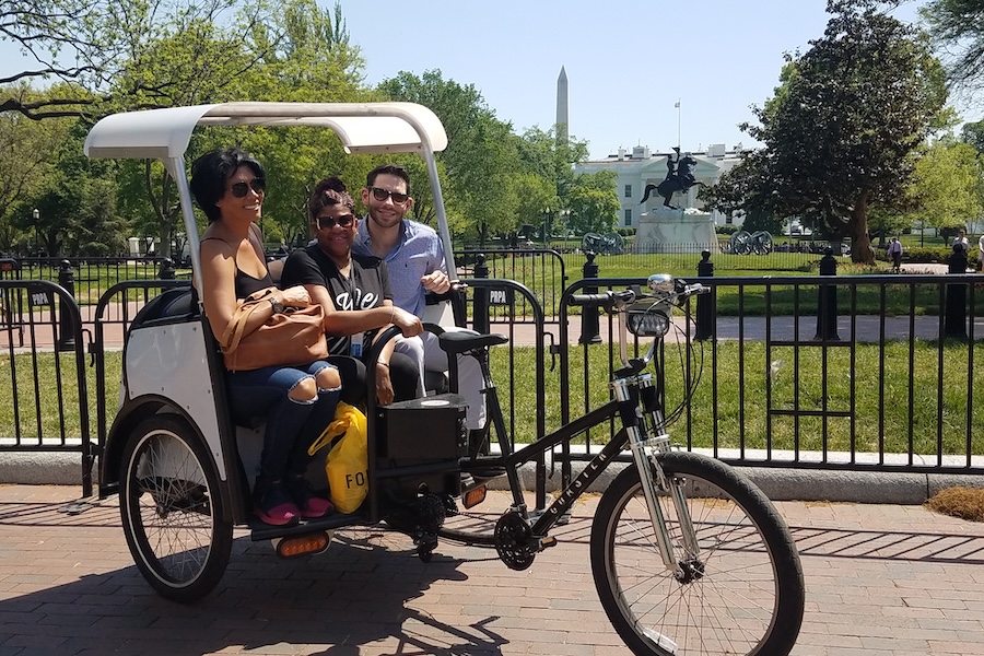Three visitors pose in a pedicab with a view of the Washington Monument in the background.