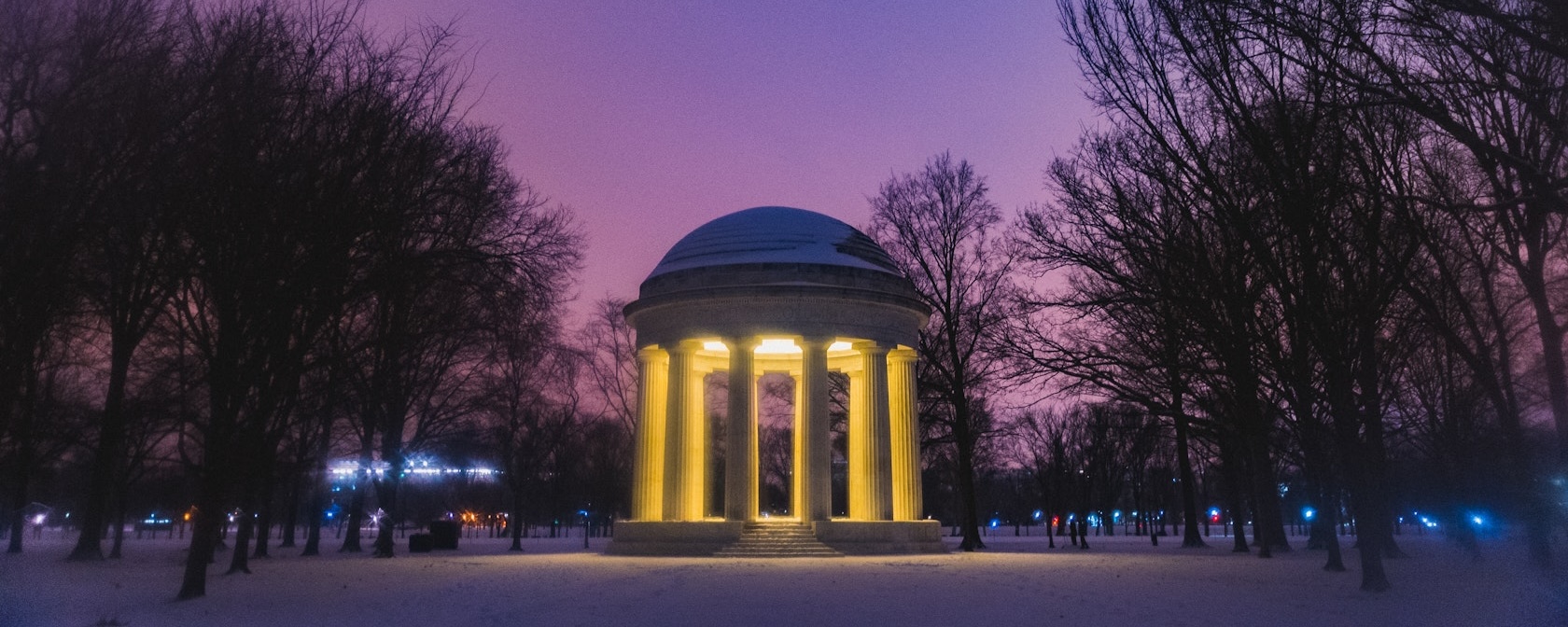 The DC War Memorial glows warmly amid snow-covered trees at twilight under a purple evening sky.