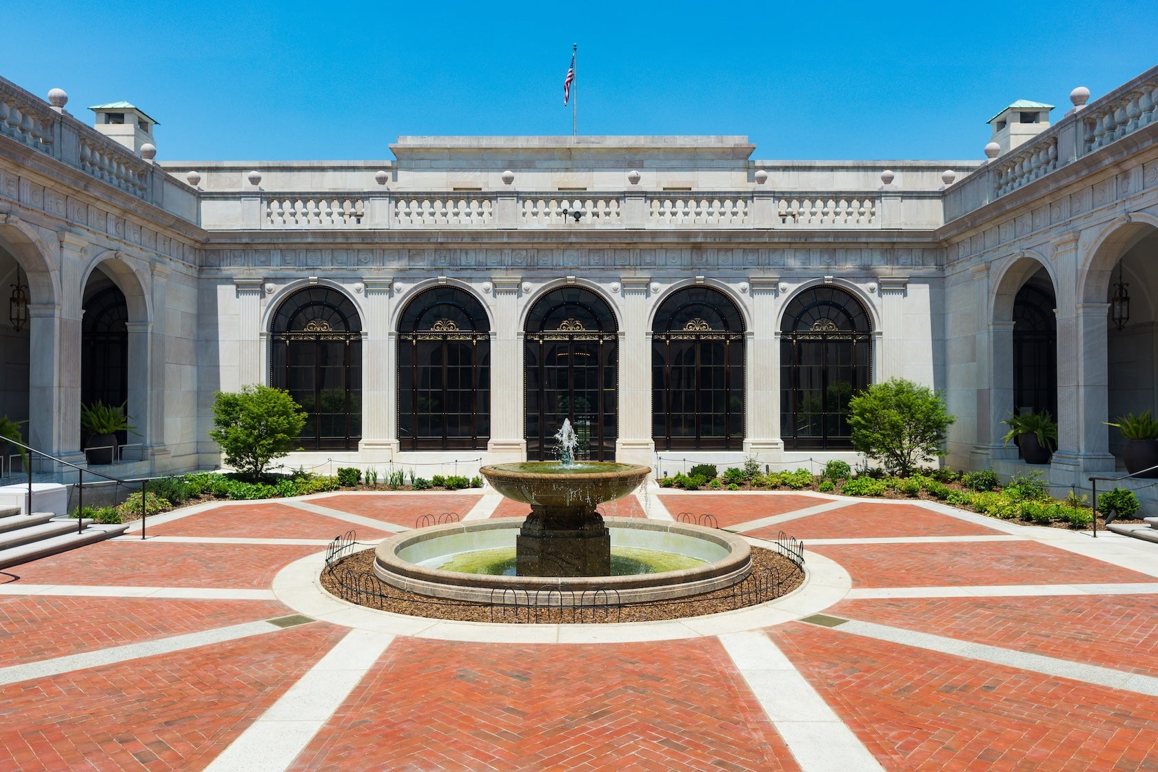 A round fountain sits at the center of a bright brick courtyard surrounded by white stone arches and large windows in Washington, DC.