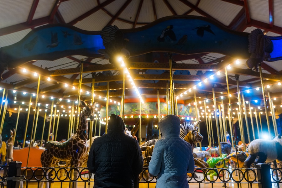 Two people in winter coats watch an illuminated carousel with animal figures during ZooLights at night.