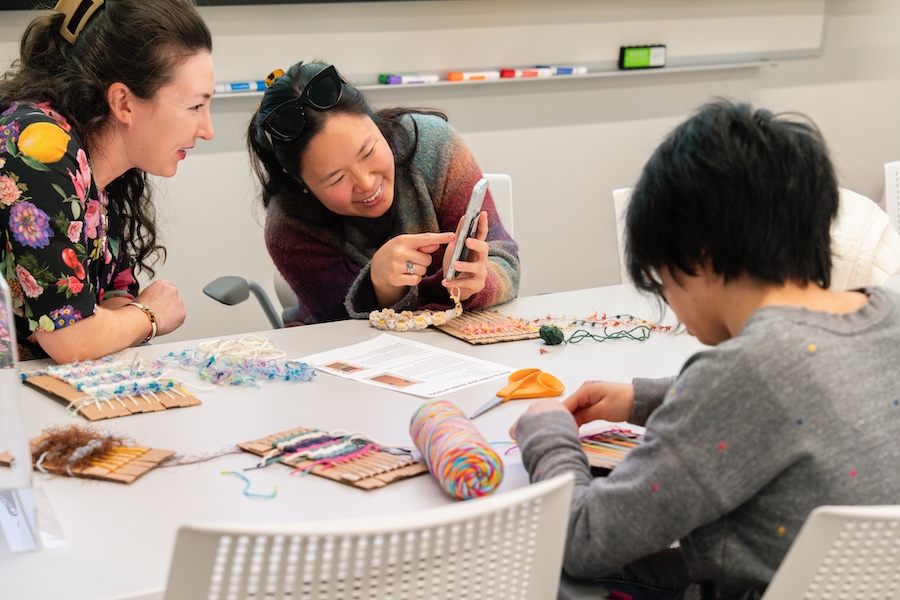 Three people sit at a table working on colorful weaving crafts; two are smiling and looking at something on a phone, while the third focuses on their weaving project.